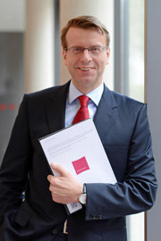 A man in a suit and red tie stands smiling by a window, holding a document in his left hand. He appears confident and professional, with glasses and a neatly styled hairstyle. The background is softly blurred, emphasizing his presence.