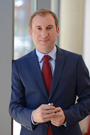 A man in a dark suit and light blue shirt stands beside a window, smiling confidently. He holds a pen in one hand, conveying a professional and approachable demeanor. The background suggests an indoor setting with natural light.