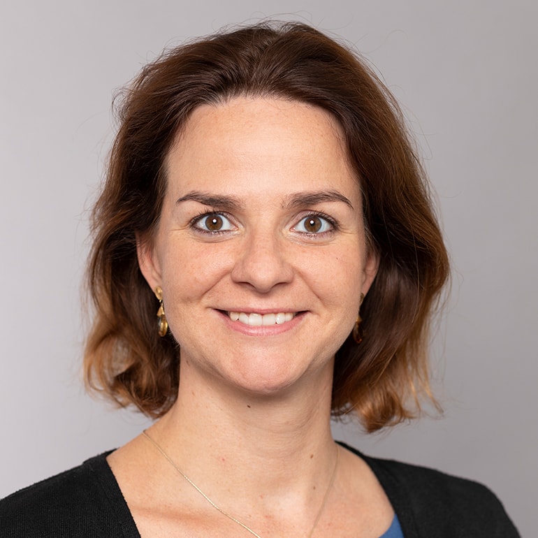 A woman with shoulder-length brown hair, wearing a black top and delicate earrings, is smiling directly at the camera. The background is a soft gray, creating a warm and approachable atmosphere.