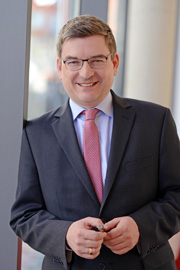 A smiling man in a business suit stands near a large window, holding a pen. He has short hair and wears glasses. The background features a blurred indoor setting, suggesting a professional environment.