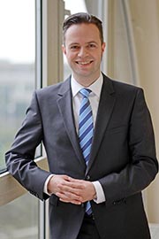 A smiling man in a suit stands confidently with his arms crossed, leaning against a window. He has short hair and is wearing a blue and white striped tie. The background suggests an office or professional setting.