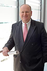 A professionally dressed man wearing a black suit and a pink tie stands smiling in an office setting, holding a rolled document. Natural light filters through large windows behind him.