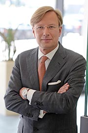 A professionally dressed man stands with arms crossed, wearing a suit and an orange tie. He has short, light-colored hair and glasses. The background suggests an indoor environment, possibly an office.