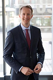 A professional man stands confidently in a suit and tie, smiling at the camera. He is positioned in front of large windows, suggesting a modern office environment. The overall impression is one of professionalism and approachability.