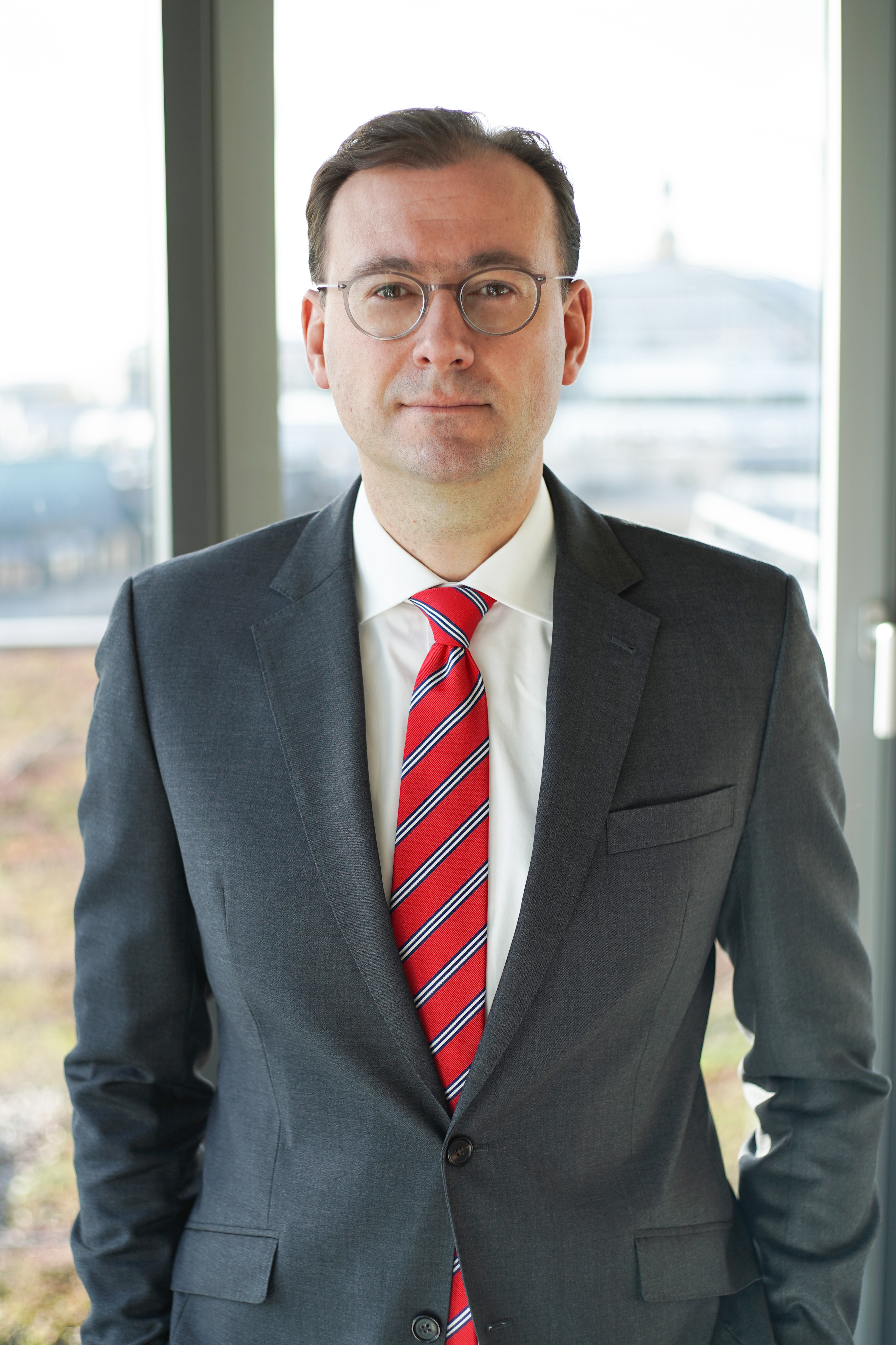 A man wearing glasses stands confidently in a dark suit and a red-striped tie, with a white shirt underneath. He is in an office setting, with large windows in the background allowing natural light to illuminate the space.