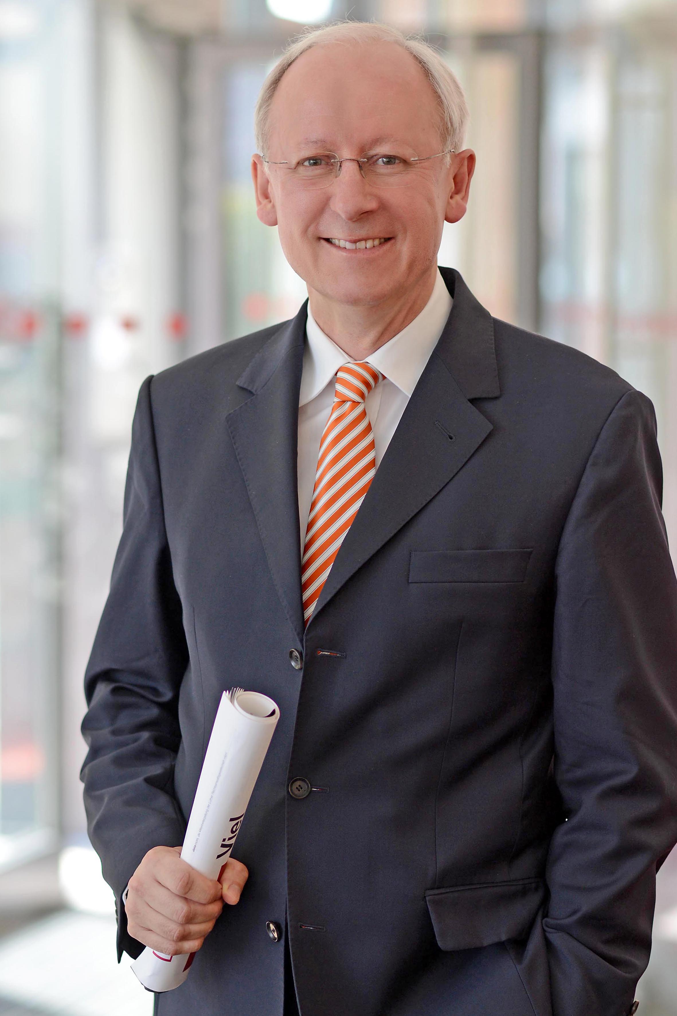 A man in a navy suit and orange-striped tie stands smiling. He holds a rolled document in one hand. The background appears to be a bright office space with glass doors.