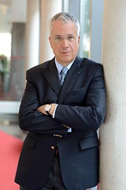 A professionally dressed man stands near a pillar, with his arms crossed. He has short gray hair and glasses, projecting a confident demeanor. The background is softly blurred, focusing attention on him.