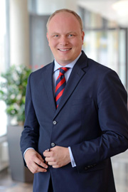 A professional-looking man is dressed in a dark suit with a red and blue striped tie. He stands confidently with his hands clasped in front of him, smiling. The background features large windows and a plant, suggesting an office setting.