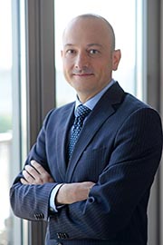 A man in a professional suit with a tie stands confidently, arms crossed, in front of a large window. He has a slight smile and a clean-shaven head, conveying a sense of approachability and professionalism. The background is softly blurred, focusing attention on him.