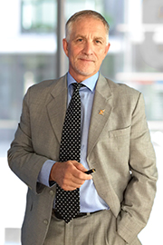A middle-aged man stands confidently, wearing a gray suit and a polka-dot tie. He holds a pen in one hand and looks directly at the camera. The background features blurred outlines of a modern office environment. His expression conveys professionalism and assurance.