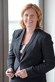 A professional woman stands confidently by a window, smiling. She wears a dark blazer over a patterned blouse. Her hair is styled in loose waves, and she holds a pen in her right hand, suggesting readiness and professionalism.