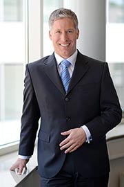 A professional man stands beside a large window, smiling warmly. He is dressed in a dark suit with a light blue shirt and tie. His posture conveys confidence and approachability. The background suggests a modern office environment.