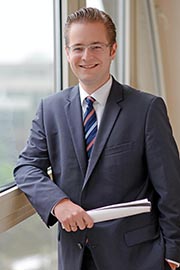 A professionally dressed man stands confidently by a window, smiling and holding a rolled document. He wears glasses and a suit with a striped tie, suggesting a business or formal context. The setting appears bright and modern.