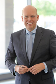 A smiling man in business attire stands confidently, holding a pen, in an office setting. He wears glasses and a light blue tie, conveying a professional demeanor. The background is bright and features large windows.
