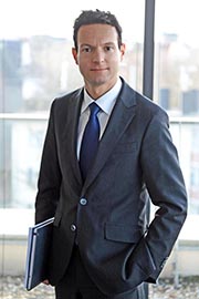 A professionally dressed man stands confidently in an office setting. He wears a dark suit and a blue tie, holding a folder in his left hand. Natural light filters through large windows behind him, creating a bright atmosphere.