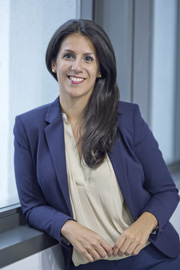 A professional woman with long dark hair smiles while leaning against a window. She wears a dark blazer over a light-colored blouse, conveying confidence and approachability in a modern office setting.