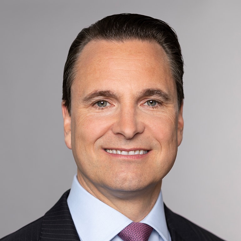 A professional man smiles confidently at the camera. He has neatly styled dark hair, light skin, and is wearing a dark suit with a light blue shirt and a patterned tie. The background is a soft gray, emphasizing his approachable demeanor.