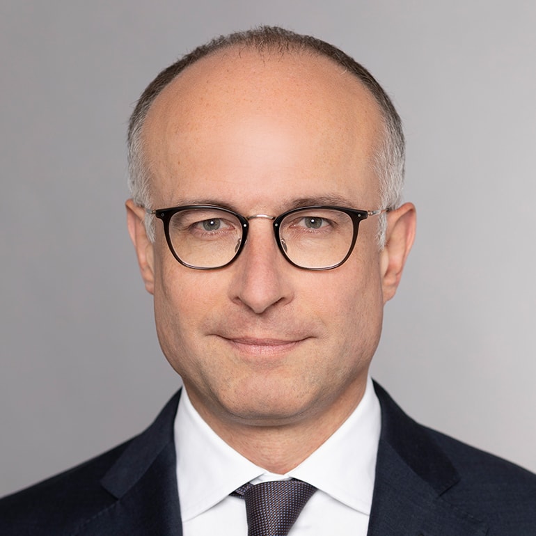 A middle-aged man with short, light hair and glasses is smiling slightly. He is wearing a dark suit and a white dress shirt with a tie. The background is a soft, neutral gray, highlighting his features.