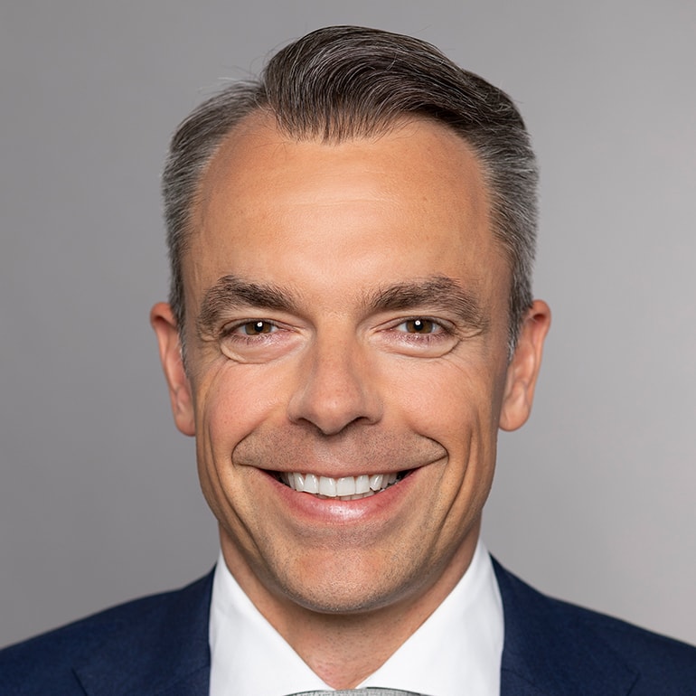 A smiling man with short, neatly styled hair is dressed in a dark suit and white shirt. He has a warm and approachable expression, with a light complexion. The background is a soft gray, highlighting his cheerful demeanor.