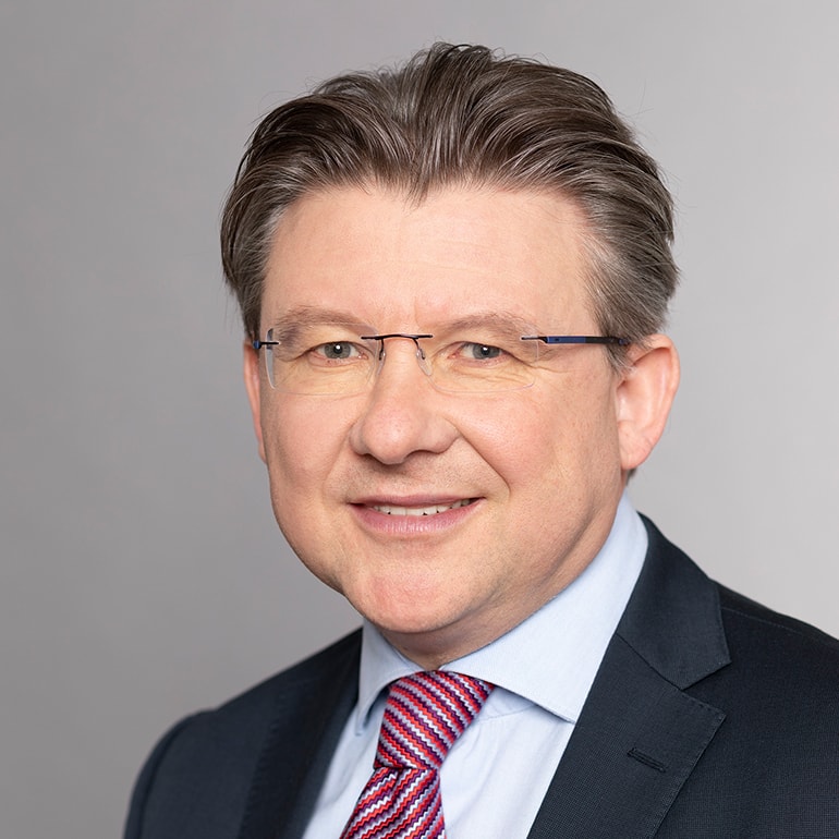 A professionally dressed man smiles at the camera. He has short, neatly styled hair and is wearing glasses. His outfit consists of a suit with a light blue shirt and a patterned tie. The background is a plain light gray.
