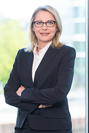 A professional woman in a dark suit stands confidently with her arms crossed. She has blonde hair and wears glasses, with a bright and modern office environment in the background.