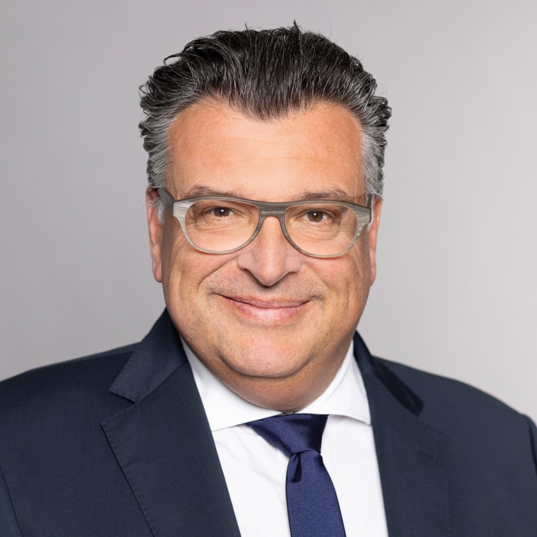 A middle-aged man with light gray hair and glasses smiles confidently. He is dressed in a dark suit with a white shirt and blue tie. The background is a plain light gray, emphasizing his professional appearance.