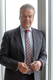 A middle-aged man in a formal suit stands confidently next to a large window, smiling. He has short, gray hair and is holding a pen in his right hand. He wears a striped tie with red and white colors.
