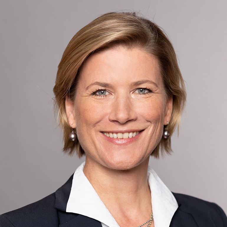 A smiling woman with short blonde hair and wearing a dark blazer over a white top. She has pearl earrings and a confident expression, set against a neutral gray background.