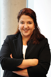 A professional woman stands confidently with her arms crossed, wearing a black blazer over a white shirt. She has dark hair styled neatly and smiles softly at the camera, set against a light background.