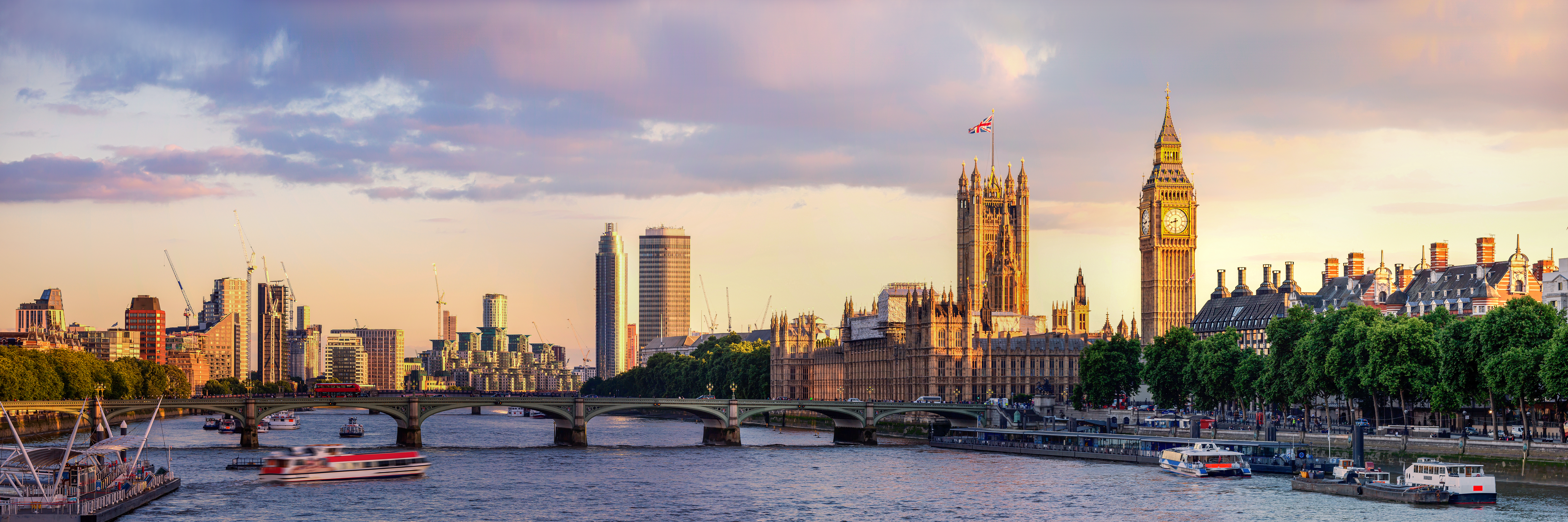 A panoramic view of London showcases the River Thames with boats, the iconic Houses of Parliament with the clock tower, and a backdrop of modern skyscrapers under a colorful sky at sunset. Lush trees line the riverbank, enhancing the cityscape's charm.