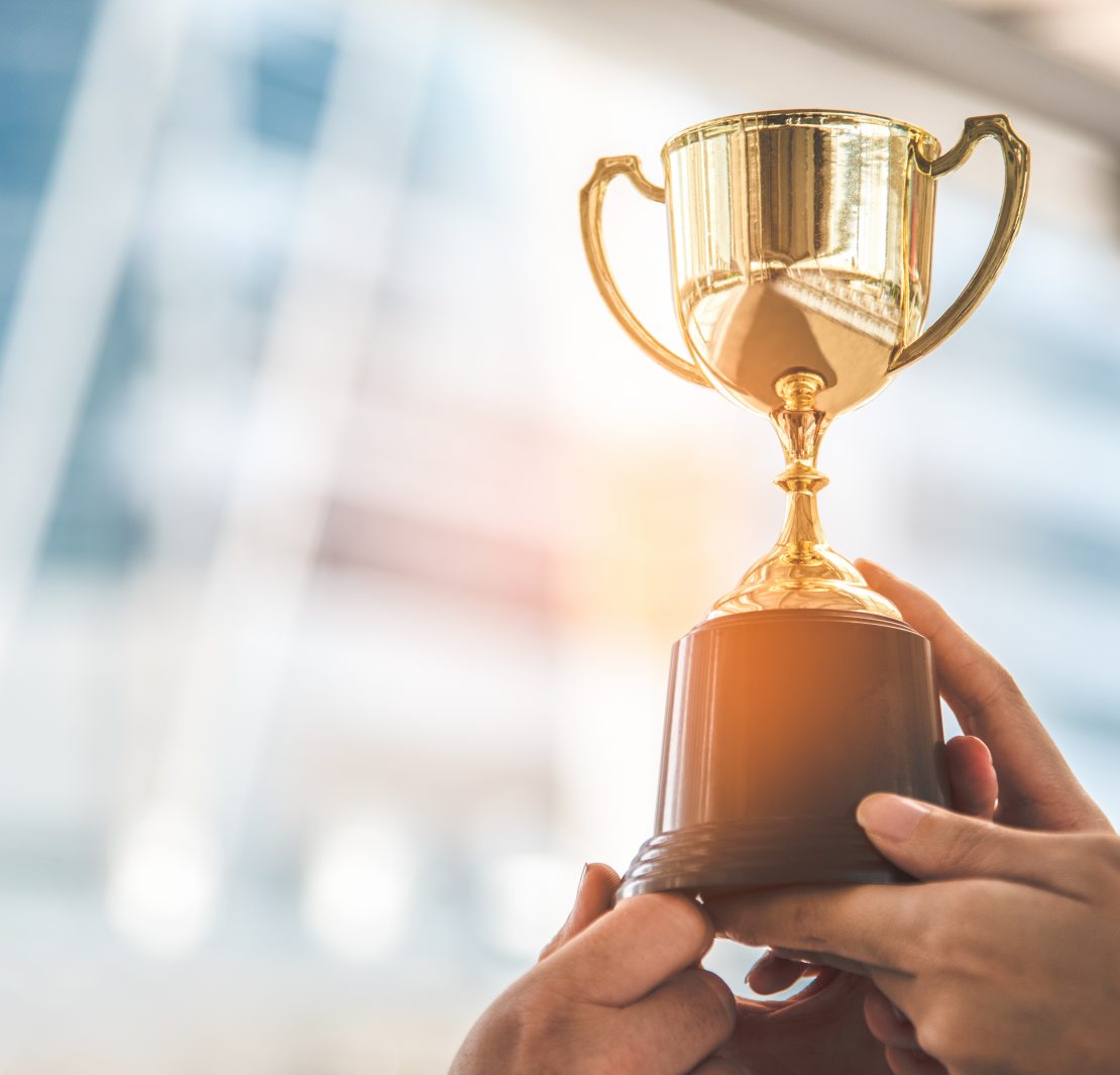 A golden trophy is held aloft by two hands, symbolizing achievement and victory. The background is blurred, emphasizing the trophy's significance in a celebratory moment.
