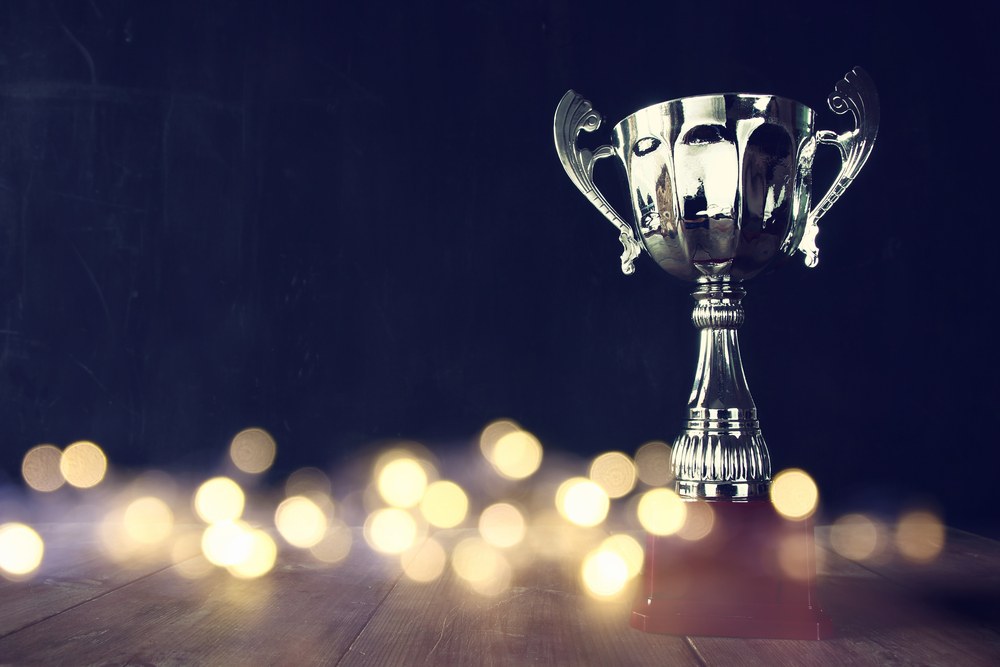 A shiny silver trophy with ornate handles stands on a dark background, symbolizing achievement and success. Soft, warm bokeh lights are blurred in the foreground, adding a celebratory atmosphere.